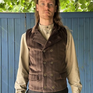 May include: A brown velvet double-breasted waistcoat featuring a subtle vertical pinstripe pattern, a notched collar, and two front flap pockets. The waistcoat is worn over a light beige long-sleeved button-down shirt with a fine vertical stripe pattern.