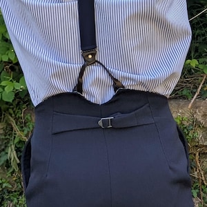 Puede incluir: Pantalones de cintura alta azul marino con trabilla y hebilla plateada. Los pantalones se combinan con una camisa abotonada a rayas azules y blancas y tirantes azul marino con detalles de cuero.