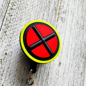 Può includere: Un portabadge circolare con un anello esterno giallo brillante e un cerchio centrale rosso diviso da una croce nera. Il dispositivo include un cordino nero retrattile, un gancio in metallo argentato e un cinturino in plastica trasparente con un bottone a pressione.