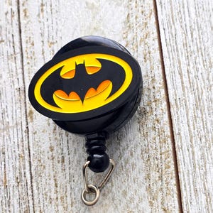 May include: A black retractable badge reel featuring a circular emblem with a yellow background and a black bat silhouette. The device includes a black plastic housing, a small black connector bead, and a silver-toned metal split ring attachment.