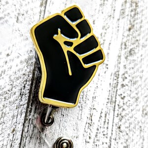 May include: A gold-colored retractable badge holder with a black raised fist design. The fist is outlined in gold.