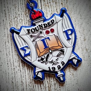 May include: A blue and white shield-shaped keychain with a detailed emblem. The emblem features a torch, open book, and Greek letters, with the word "FOUNDED" and the year "1922" visible. The keychain has a blue border and a silver keyring.