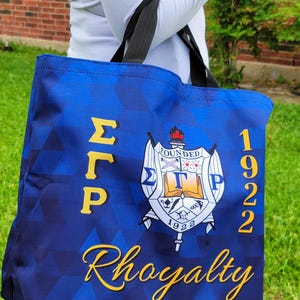 May include: Blue tote bag with a gold Greek letter design, a crest with the letters "ΣΓΡ" and the text "Founded 1922", and the word "Rhogalty" in gold script.