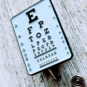 May include: A white eye chart with black letters on a black and silver retractable badge holder. The eye chart has the letters E, FP, TOZ, LPED, PECFD, EDFCZP, FELOPZD, and DEFPOTEC. The numbers 1 through 8 are listed on the right side of the chart.