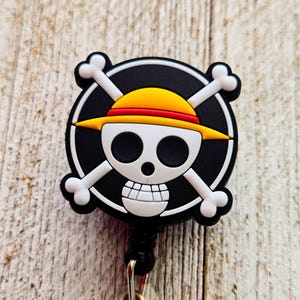 May include: A circular badge reel featuring the One Piece skull and crossbones logo. The design includes a white skull wearing a yellow straw hat with a red band, set against a black circular background with white crossbones extending outward.
