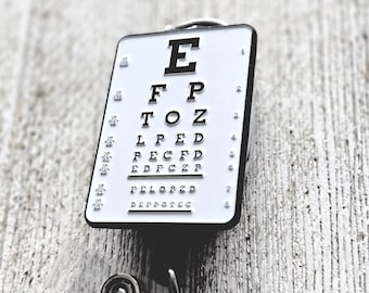 May include: A rectangular white badge reel featuring a black Snellen eye chart design with the letters E, F P, T O Z, L P E D, P E C F D, E D F C Z P, F E L O P Z D, and D E P R O T E C, along with numerical visual acuity fractions on the sides.