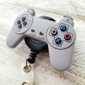 Puede incluir: Un llavero de plástico gris y negro en forma de mando de videojuego con un cordón retráctil. El mando tiene las palabras "Select" y "Start" impresas. El mando tiene una disposición de botones en blanco y negro con botones de colores.