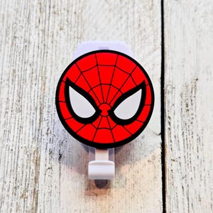 May include: A white and red Spider-Man themed car visor clip. The clip features a red circle with a black spiderweb design and white eye shapes. The clip is made of plastic and is designed to attach to a car visor.