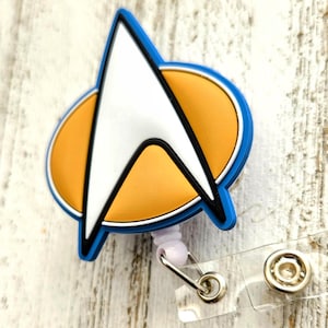 Op de afbeelding: Een Star Trek commandodivisie-badge in de vorm van een deltashild met een wit midden, zwarte omlijning en een gouden ovale achtergrond. De badge is bevestigd aan een uittrekbare kunststof haspel met een doorzichtige vinylband en zilveren drukknoop.