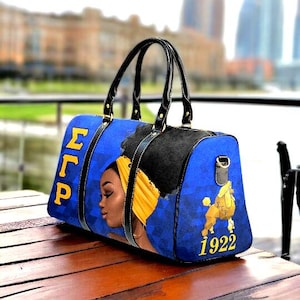 May include: Blue and black travel duffel bag with a portrait of a woman with a yellow head wrap and a poodle. The bag has the letters "ΣΓΡ" and the number "1922" printed on it.