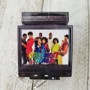 May include: A small television ornament with a retro design, featuring the cast of "A Different World." The TV has a black frame and a VCR on top. The show's title is displayed in colourful text on the screen.