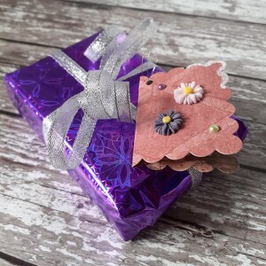 May include: A purple gift box with a silver ribbon and a pink paper tag with a flower design. The tag has a scalloped edge and is attached to the box with a silver string.