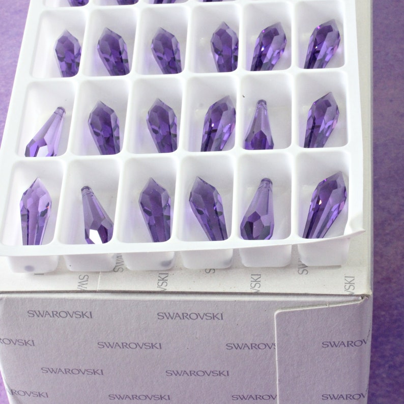 May include: A box of 24 purple crystal teardrop pendants, arranged in a grid pattern. The box is labeled "Swarovski" on the front and sides.