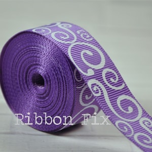 May include: A roll of purple ribbon with a white scroll pattern. The ribbon has a ribbed texture and the text "Ribbon Fix" is visible at the bottom. This ribbon is suitable for crafts and gift wrapping.