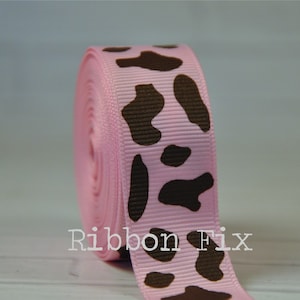 Peut inclure: Rouleau de ruban gros-grain rose avec un motif répété de taches de vache marron. Le texte "Ribbon Fix" est imprimé en blanc sur le ruban. Le ruban est enroulé serré, montrant la texture et les variations de couleur.