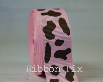 May include: A roll of pink grosgrain ribbon with a repeating pattern of brown cow spots. The text "Ribbon Fix" is printed in white on the ribbon. The ribbon is tightly wound, showing the texture and color variations.