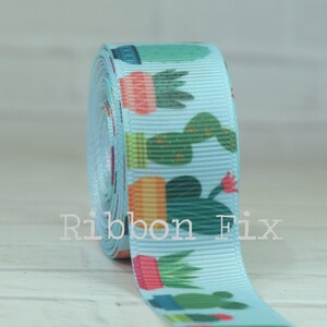 May include: A roll of light blue ribbon with a repeating pattern of potted cacti in various colors. The ribbon has a ribbed texture and the text "Ribbon Fix" is printed on the ribbon.