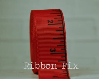 May include: A roll of red measuring tape ribbon with black markings. The ribbon has a ruler design printed on the edge, with numbers and lines indicating inches. The text "Ribbon Fix" is printed on the ribbon.