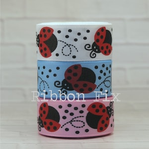 Può includere: Tre rotoli di nastro impilati, bianco, azzurro e rosa. Ogni nastro presenta un motivo ripetuto di coccinelle rosse e nere con pois neri e linee a spirale. Il testo "Ribbon Fix" è stampato sui nastri.
