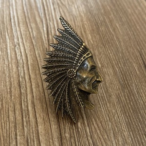 May include: A gold-toned metal pin featuring a detailed profile of a Native American man wearing a headdress with feathers. The pin is a decorative piece with a vintage aesthetic.