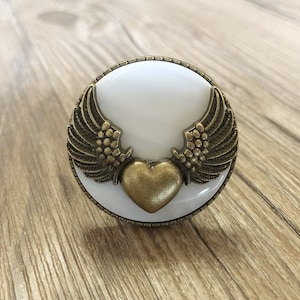 Peut inclure: Un bouton en céramique blanche avec un cœur et des ailes dorés. Le bouton est rond et a une surface lisse.