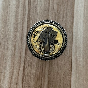 May include: A round, gold and black metal bottle stopper with a steampunk design. The stopper features a gear-like design with playing cards and a bottle opener.