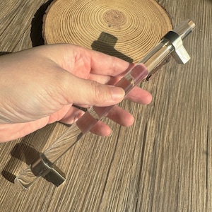 以下が含まれることがあります： シルバーの金属製ハンドルとピンクとクリアのチェック柄が付いた、透明なガラス製のボトルストッパー。木製の背景に、手でストッパーを持っています。