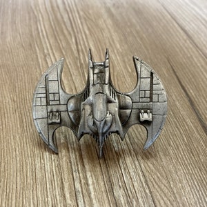 Op de afbeelding: Een zilveren metalen pin in de vorm van een vleermuis met een ruimteschipontwerp. De vleermuis heeft een gedetailleerd ontwerp met een textuur.