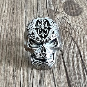 May include: A silver skull ring with intricate floral designs. The skull has a detailed face with a cross design on the forehead.