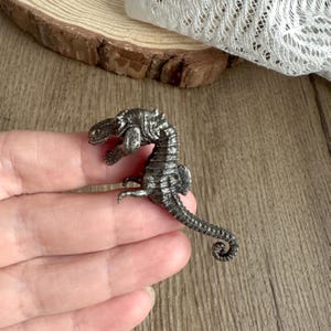 Pode incluir: Uma figura detalhada de cavalo-marinho prateado. O cavalo-marinho é intrincadamente projetado com escamas texturizadas e uma cauda enrolada. A figura é segurada em uma mão, mostrando seu tamanho e detalhes. O fundo inclui uma superfície de madeira e um pedaço de renda.