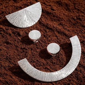 May include: A set of four silver-toned metal hardware pieces featuring a textured, sunburst-like pattern. The collection includes one semi-circular handle, one large circular knob, one smaller circular knob, and one curved, crescent-shaped handle.