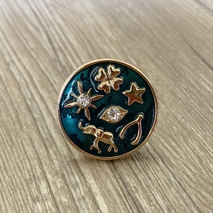 Puede incluir: Un anillo de tono dorado con una base de esmalte verde. El anillo presenta una variedad de encantos, incluyendo un trébol de cuatro hojas, una estrella, un sol, un elefante, una herradura y un mal de ojo.