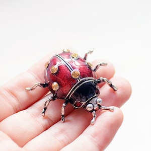 Könnte beinhalten: Eine rote und schwarze Marienkäfer-Brosche mit goldfarbenen Akzenten und Strasssteinen. Die Brosche ist aus Metall und hat ein detailliertes Design.