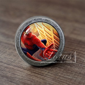 May include: A silver-toned round cabinet knob with a Spiderman image. The image shows Spiderman in a red and blue suit, hanging from a building. The background is a yellow and orange brick wall.