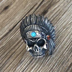 Puede incluir: Un anillo de calavera de plata con una piedra turquesa y piedras naranjas, que lleva un tocado con plumas.
