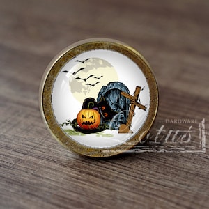 Pode incluir: Um botão de armário redondo de cor dourada com um inserto de cerâmica branca que apresenta um design temático de Halloween. O design inclui uma abóbora de Halloween, um gato preto, uma lápida e morcegos voando na frente de uma lua cheia.