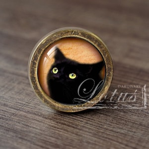 May include: A black cat with yellow eyes is featured in a round, antique gold-toned cabinet knob. The cat is looking to the left of the image.