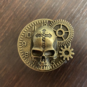 May include: A gold-toned metal skull with a cross on its forehead, surrounded by gears and a clock face with Roman numerals.
