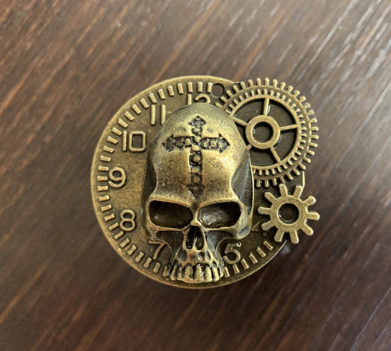 May include: A gold-toned metal skull with a cross on its forehead, surrounded by gears and a clock face with Roman numerals.