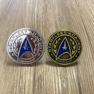 Peut inclure: Deux épingles de commande de l'Académie de la flotte stellaire. L'une est argentée avec un emblème bleu et argent. L'autre est dorée avec un emblème bleu et or.
