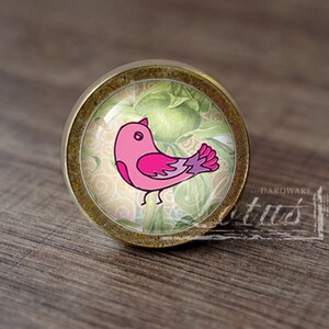 May include: A round, bronze-colored cabinet knob with a glass insert featuring a pink bird with a purple tail on a green and yellow floral background.