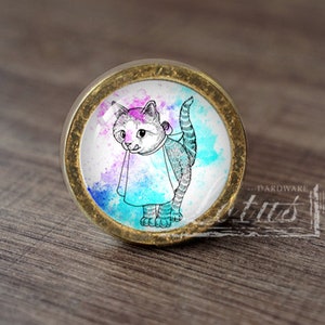 以下が含まれることがあります： 水彩画風のデザインが施されたガラス製のインサートがついた、丸い金色のキャビネットノブ。青い紫色のマントを着た猫が描かれています。猫は黒で縁取られ、白い背景になっています。