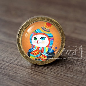May include: Circular cabinet knob with a bronze finish. It features a colourful illustration of a white cat wearing a winter hat, scarf, and mittens. The cat has one green and one blue eye. The background is orange.