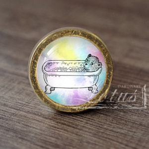 以下が含まれることがあります： 水彩画のようなカラフルな背景を持つ、丸い金色のキャビネットノブ。ノブには、バスタブで入浴するクマの白黒のイラストが描かれています。