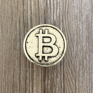 Könnte beinhalten: Eine goldfarbene Metallmünze mit einem erhabenen, stilisierten "B" in der Mitte, das die Kryptowährung Bitcoin darstellt.