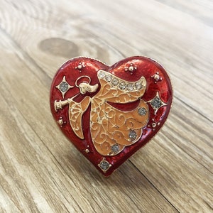 May include: A red heart-shaped trinket box featuring an angel playing a trumpet. The angel is gold with a red and gold design. The box is decorated with rhinestones.
