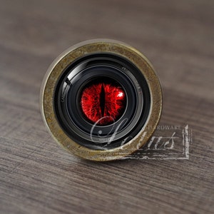 Puede incluir: Primer plano de un objetivo de cámara con un ojo rojo en el centro. El objetivo está rodeado por un anillo de color latón.