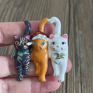 May include: A trio of enamel cat figurines, each with a different colour and design. The black cat is wearing a green scarf, the orange cat has a crown of rhinestones, and the white cat has a green holly sprig.