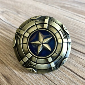 Może przedstawiać: Złota klamra do paska z niebieskim i złotym wzorem gwiazdy. Klamra jest okrągła i ma fakturowaną powierzchnię.