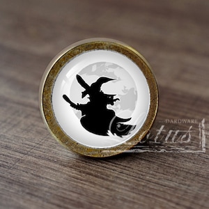 Pode incluir: Um botão de armário redondo, de cor bronze, com fundo branco e uma silhueta preta de uma bruxa voando em uma vassoura na frente da lua cheia.
